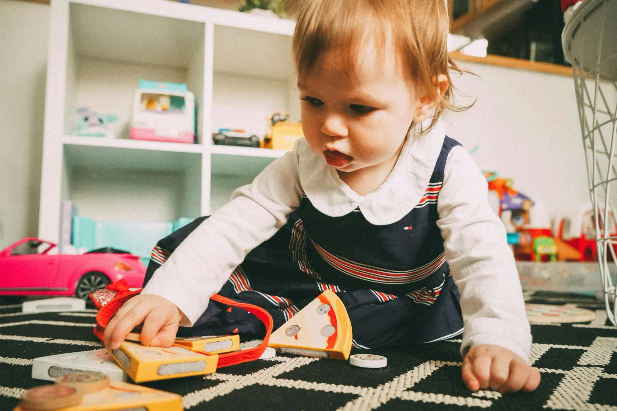 A toddler in a striped dress plays with wooden toy pizza slices on a patterned rug